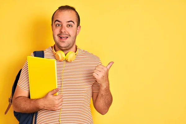 バックパックのヘッドフォンを身に着けている学生の男は 孤立した黄色の背景を指している上にノートブックを保持し 幸せな顔笑顔で親指で側面まで示す — ストック写真