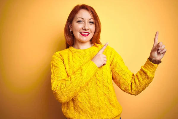 美丽的红头发女人穿着冬季毛衣 站在孤立的黄色背景上微笑着 看着摄像机 用双手和手指指向旁边 — 图库照片
