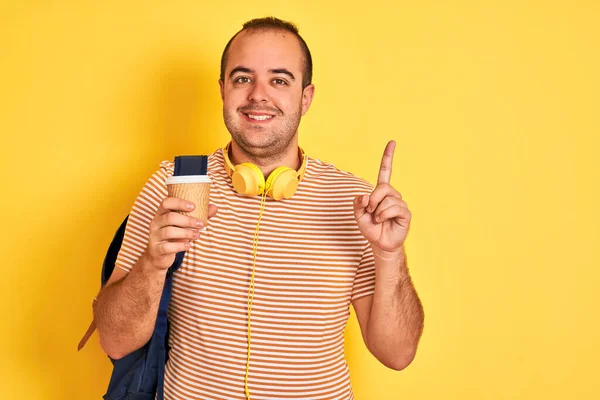 バックパックを身に着けている学生の男は幸せな顔でアイデアや質問指差す指で驚いて隔離された黄色の背景にコーヒーを飲む ナンバーワン — ストック写真