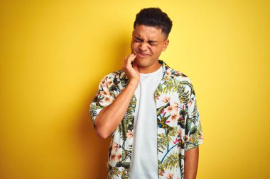 Tatilde olan Brezilyalı genç bir adam, izole edilmiş sarı arka plan üzerine çiçekli tişört giyerek diş ağrısı ya da diş ağrısı yüzünden ağız ve acı dolu bir ifadeyle dokunuyor. Dişçi konsepti.