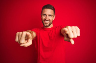 Kırmızı izole edilmiş arka plan üzerine gündelik tişört giyen genç yakışıklı adam seni ve kamerayı parmaklarıyla gösteriyor, gülümsüyor ve neşeli.