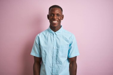 Yüzünde mutlu ve havalı bir gülümseme olan, izole edilmiş pembe arka planda duran mavi günlük gömlekli Afro-Amerikan bir adam. Şanslı kişi..