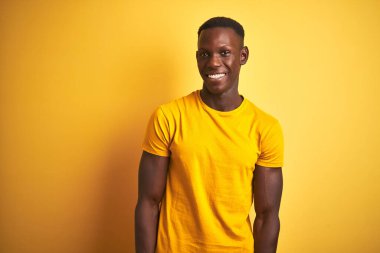 Sıradan tişörtler giyen genç Afro-Amerikan bir adam izole edilmiş sarı arka planda dikilip yüzünde gülümseme, doğal bir ifade ile bir o yana bir bu yana bakıyor. Kendinden emin gülen.