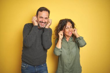 Yüksek sesle müzik sesi için rahatsız ifade ile parmakları ile kulakları kapsayan izole sarı arka plan üzerinde Güzel orta yaş çift. Sağır kavramı.