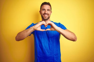 Sarı izole edilmiş arka plan üzerine sıradan mavi tişörtler giyen genç yakışıklı bir adam. Aşkla gülümsüyor. Elleriyle kalp sembolü ve şekli gösteriyor. Romantik konsept.