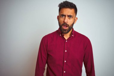 Kırmızı şık gömlek giyen genç Hintli bir adam. Gri arka planda duruyor. Şok olmuş yüz ifadesiyle, şüpheci ve alaycı görünüyor.