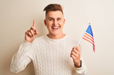 İzole edilmiş beyaz arka plan üzerinde Usa bayrağı tutan genç yakışıklı bir adam bir fikir veya soru işareti ile şaşırdı mutlu bir yüzle, bir numara