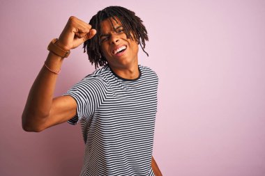 Rasta desenli tişörtlü Afro adam izole edilmiş pembe arka planda dikiliyor kızgın ve öfkeyle bağırırken yumruğunu kaldırıyor. Öfke ve agresif kavram.