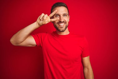 Genç yakışıklı adam kırmızı izole arka plan üzerinde rahat t-shirt giyen yüzü üzerinde parmakları ile barış sembolü yapıyor, zafer gösteren neşeli gülümseyen