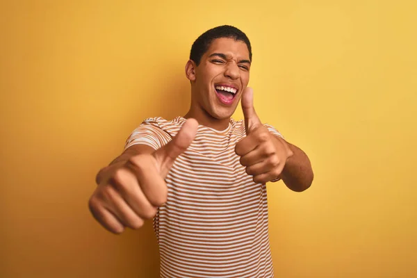 年轻英俊的阿拉伯男子 身穿条纹T恤 站在孤立的黄色背景上 赞许用手做积极的手势 微笑着竖起大拇指 为成功感到高兴 赢的手势 — 图库照片
