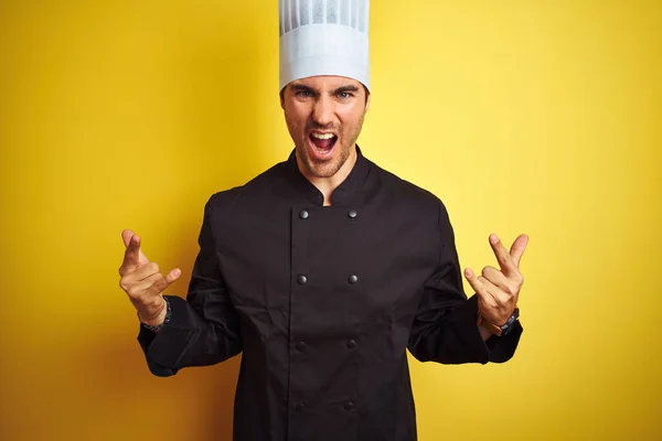 年轻厨师 身穿制服 头戴帽子 站在孤立的黄色背景之上 用疯狂的表情高举双手做着摇滚符号 音乐明星 沉重的概念 — 图库照片