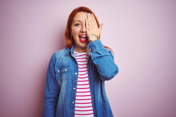 Beautiful redhead woman wearing denim shirt and striped t-shirt over isolated pink background covering one eye with hand, confident smile on face and surprise emotion.