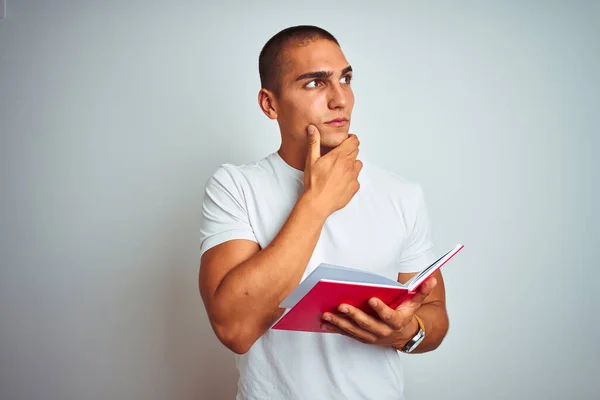 Young handsome man reading red book over white isolated background serious face thinking about question, very confused idea