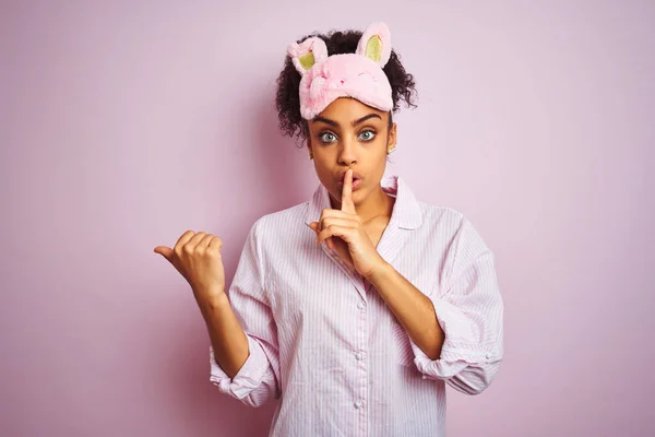 年轻的非洲裔美国女人 戴着睡衣和面罩 披着孤立的粉色背景 要求保持安静 手指放在嘴唇上 手拉手指向侧面 沉默和秘密概念 — 图库照片