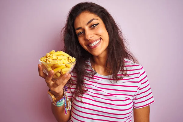 Young beautiful woman holding bowl with dry macaroni pasta over isolated pink background with a happy face standing and smiling with a confident smile showing teeth