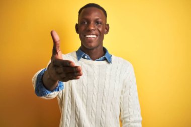 İzole edilmiş sarı arka planda beyaz kazak ve kot gömlek giyen Afrikalı Amerikalı bir adam selamlama ve hoş geldin demek için gülümseyerek tokalaşıyor. Başarılı bir iş..