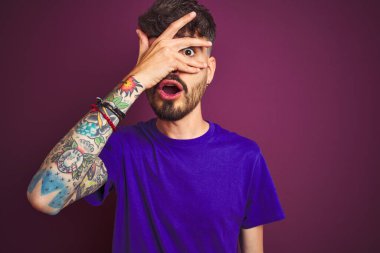 İzole edilmiş mor arka plan üzerinde duran dövmeli genç bir adam. Şok içinde, yüzünü ve gözlerini kapayarak utangaç ifadesiyle parmaklarının arasından bakıyor..
