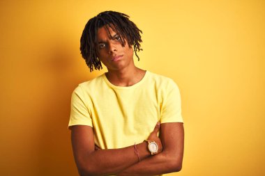 Rastgele tişörtler giyen Afro bir adam izole edilmiş sarı arka plan üzerinde duruyor şüpheci ve gergin, yüzünde çapraz kollu tasvip etmeyen bir ifade var. Negatif kişi.