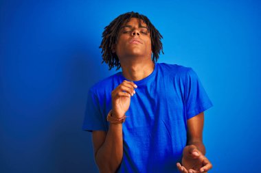 Rastalı tişörtlü Afro-Amerikan bir adam mavi arka plan üzerinde dikiliyor tiksindirici ve tiksindirici bir yüz ifadesi takınıyor çünkü tiksinti tepkisi veriyor. Eller havada. Can sıkıcı bir kavram..