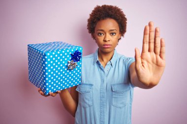 Afrika kökenli Amerikalı bir kadın pembe izole edilmiş arka planda doğum günü hediyesi tutuyor. Açık elle dur işareti yapıyor. Ciddi ve kendinden emin bir ifadeyle, savunma jesti.