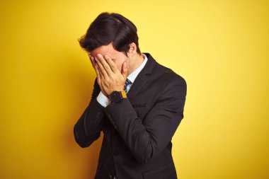 Takım elbiseli ve kravatlı genç yakışıklı bir işadamı, izole edilmiş sarı arka planda dikiliyor ve ağlarken suratını ellerle kapatıyor. Depresyon kavramı.