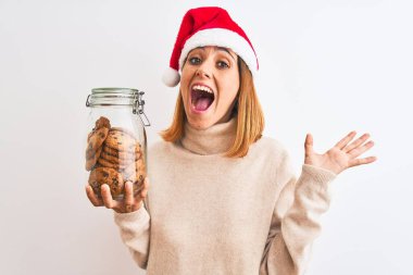 Güzel kızıl saçlı kadın, elinde kurabiye kavanozuyla izole edilmiş arka planda mutlu ve heyecanlı, kazanan yüz ifadesi zaferi büyük bir gülümsemeyle kutluyor ve ellerini kaldırıyor.