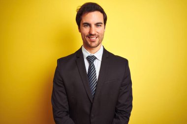 Takım elbiseli ve kravatlı genç yakışıklı işadamı mutlu ve havalı bir gülümsemeyle izole edilmiş sarı arka planda duruyor. Şanslı kişi..