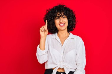 Kıvırcık saçlı, beyaz tişörtlü, izole edilmiş kırmızı arka planı olan genç bir Arap kadın bir numarayı işaret ederken kendinden emin ve mutlu bir şekilde gülümsüyor..