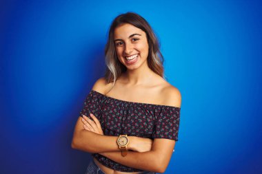 Mavi izole edilmiş arka planda çiçekli tişört giyen güzel bir kadın kollarını kavuşturup kameraya bakarken gülümsüyor. Pozitif kişi.