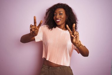 Genç Afro-Amerikan kadın tişört giyip pembe arka planda dikiliyor. Kameraya bakıp zafer işareti yapıyor. İki numara..