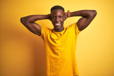 Genç Afro-Amerikalı bir adam günlük tişörtler giyiyor. İzole edilmiş sarı arka planda duruyor. Kolları ve elleri başının arkasında, mutlu bir şekilde gülümsüyor.