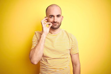 Sakallı, sarı izole edilmiş arka plan ağzı ve dudakları parmaklarıyla kapanmış fermuarı olan sıradan çizgili tişörtlü genç kel bir adam. Gizli ve sessiz, tabu konuşması
