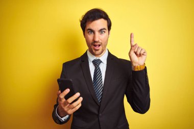 Genç yakışıklı iş adamı, izole edilmiş sarı arka plan üzerinde duran akıllı telefonu kullanarak bir fikir veya soru işareti ile şaşırdı.