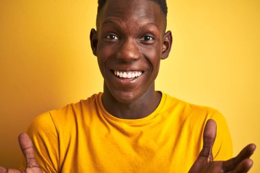 Siyah tişörtlü Afro-Amerikalı adam izole edilmiş sarı arka planda dikiliyor mutlu ve heyecanlı, kazanan yüz ifadesi zaferi büyük bir gülümsemeyle kutluyor ve ellerini kaldırıyor.