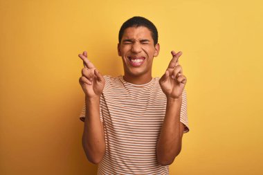 İzole edilmiş sarı arka plan üzerinde duran çizgili tişört giyen genç, yakışıklı Arap adam umut ve gözler kapalı bir şekilde gülümseyerek işaret ediyor. Şans ve batıl inanç..