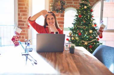 Masada oturan güzel bir kadın evde dizüstü bilgisayarla çalışıyor Noel ağacının etrafında gülümsüyor elleri ve parmakları gülen yüzlü bir çerçeve yapıyor. Yaratıcılık ve fotoğrafçılık kavramı.