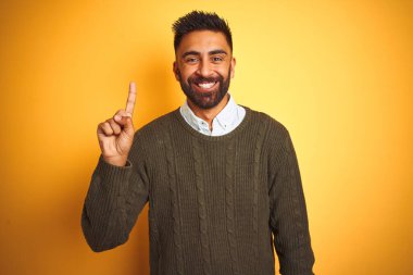 Yeşil kazak ve tişört giyen genç Hintli adam izole edilmiş sarı arka plan üzerinde duruyor ve bir numaralı parmağını gösterirken kendinden emin ve mutlu bir şekilde gülümsüyor..