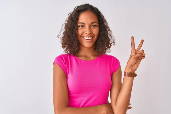ピンクのTシャツを着た若いブラジル人女性が白い背景の上に立っていて カメラが勝利のサインをして幸せそうな顔をして笑っています 第二番 — ストック写真