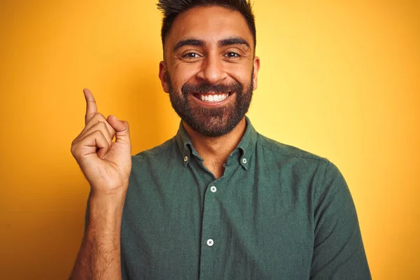カメラを見ている側に手と指で指して 顔に大きな笑顔で隔離された黄色の背景の上に立って緑のシャツを着て若いインドの男 — ストック写真