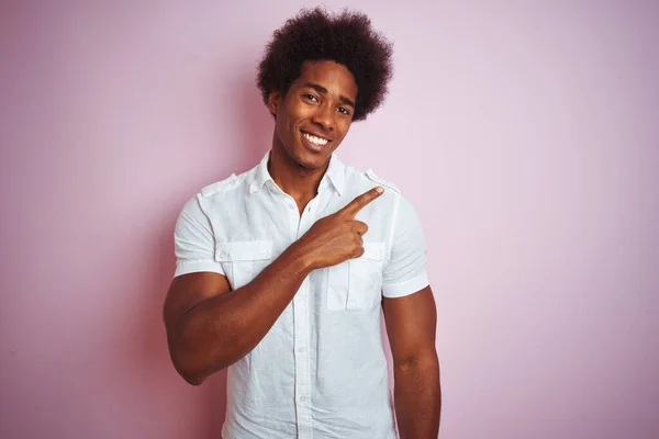 白いシャツを着たアフロの髪をした若いアメリカ人男性は 顔の笑顔で陽気にピンクの背景に立って手と指で顔に幸せと自然な表情で側面まで指しています — ストック写真
