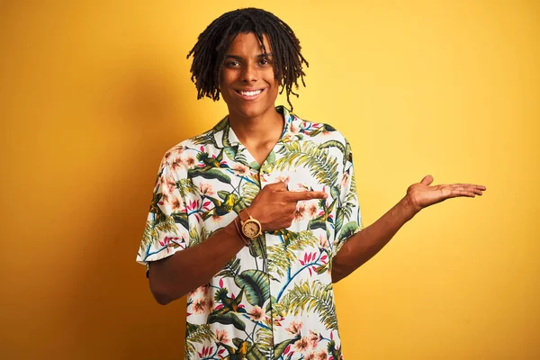 Afro man with dreadlocks on vacation wearing summer shirt over isolated yellow background amazed and smiling to the camera while presenting with hand and pointing with finger.