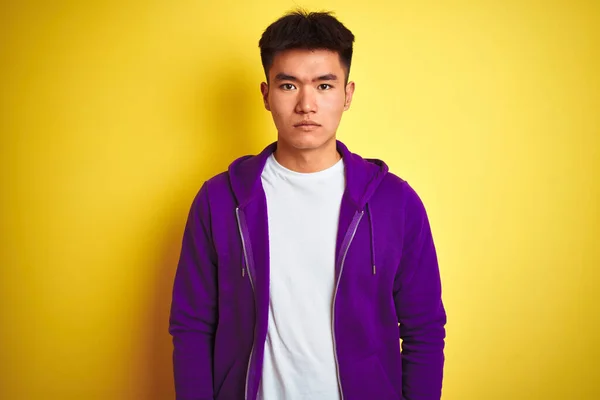 孤立した黄色の背景の上に紫色のスウェットシャツを着た若いアジア系中国人男性が顔に深刻な表情でリラックスしています シンプルで自然なカメラを見て — ストック写真