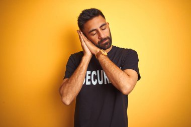 Arap Hindistanlı İspanyol güvenlik üniforması giyen izole edilmiş sarı arka planda uyuyan yorgun ve elleri ile birlikte poz veren kapalı gözlerle gülümseyen adam..