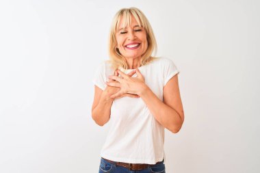 Orta yaşlı bir kadın, izole edilmiş beyaz arka plan üzerinde sıradan bir tişört giyer, elleri göğsünde, gözleri kapalı ve yüzünde minnet dolu bir jest ile gülümser. Sağlık kavramı.