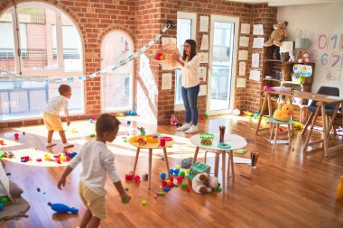 Anaokulunda bir sürü oyuncağın etrafında top ve hasır sepet kullanarak basketbol oynayan güzel öğretmenler ve yeni yürümeye başlayan çocuklar.