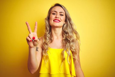 Sarı arka plan üzerinde duran genç saldırgan kadın kendini beğenmiş ve mutlu bir şekilde gülümserken iki numaralı parmaklarıyla işaret ediyor..
