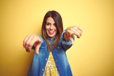 Sarı arka planda duran genç ve güzel bir kadın seni ve kamerayı işaret ediyor, pozitif ve neşeli gülümsüyor.