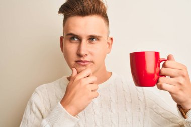 Kırmızı kahve içen yakışıklı genç adam izole edilmiş beyaz arka planda duruyor. Ciddi bir yüz. Soru soruyor, kafası karışık bir fikir.