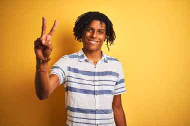 Sarı arka plan üzerinde çizgili gömlek giyen Afro-Amerikalı bir adam iki numarayı gösterirken kendinden emin ve mutlu bir şekilde gülümsüyor..