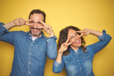 Güzel orta yaş çift birlikte izole sarı arka plan üzerinde ayakta yüz üzerinde parmaklar ile Barış sembolü yapıyor, neşeli gösteren zaferle gülümseyen